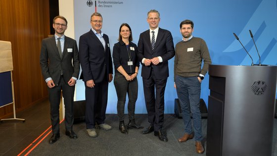 Eine Frau im Business Look steht zwischen vier Männern. Alle schauen in die Kamera